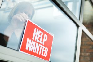 Sign of hiring, recruitment, employment and economic recovery through a woman business person posting a "HELP WANTED" sign for new employees.